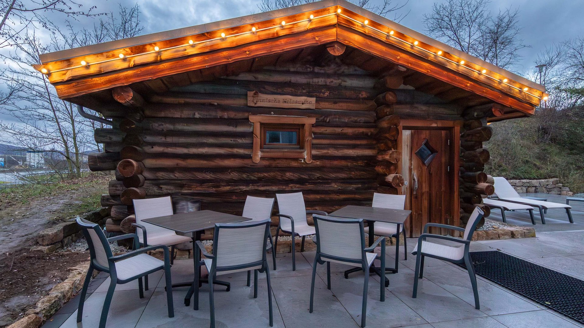 Wooden cabin with string lights, outdoor area with tables and chairs in the evening