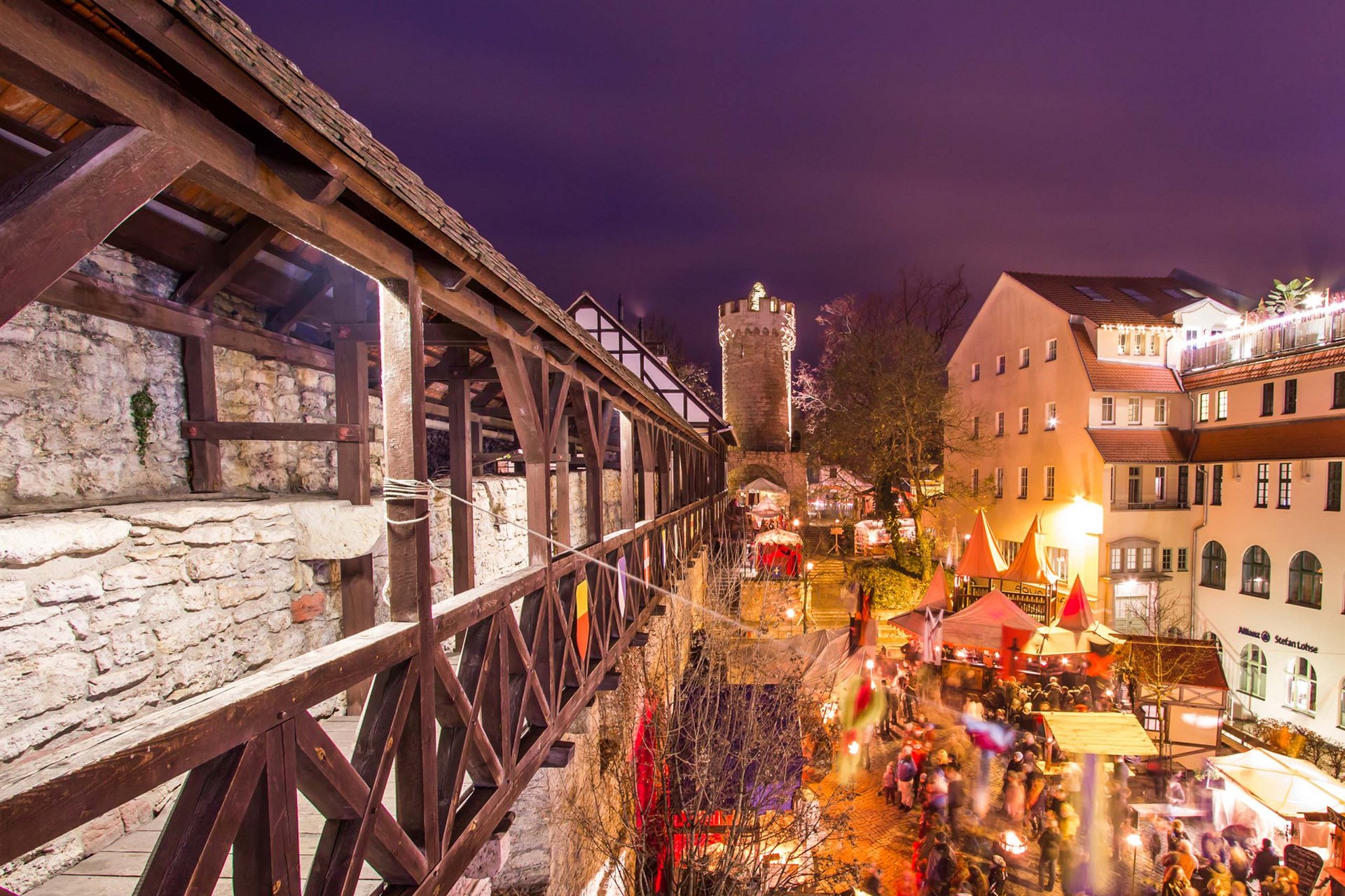 Beleuchteter mittelalterlicher Markt mit Menschen neben alter Stadtmauer und Turm bei Nacht
