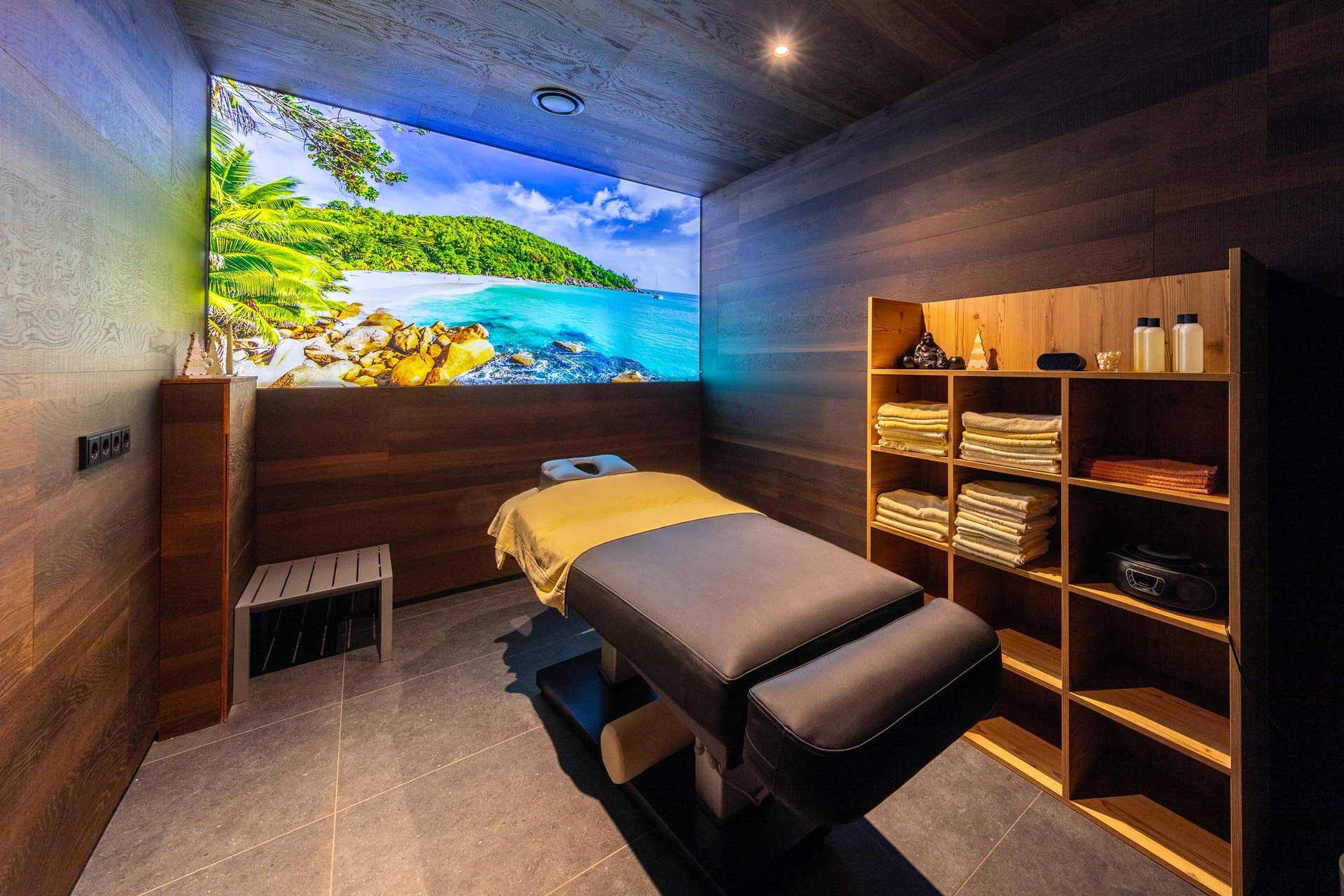 Moderne Massageraum mit tropischem Strandbild und Holzregal mit Handtüchern