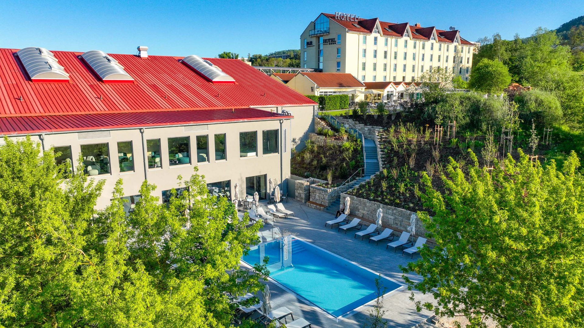 Hotel complex with outdoor pool, lounge chairs, and greenery under clear blue sky