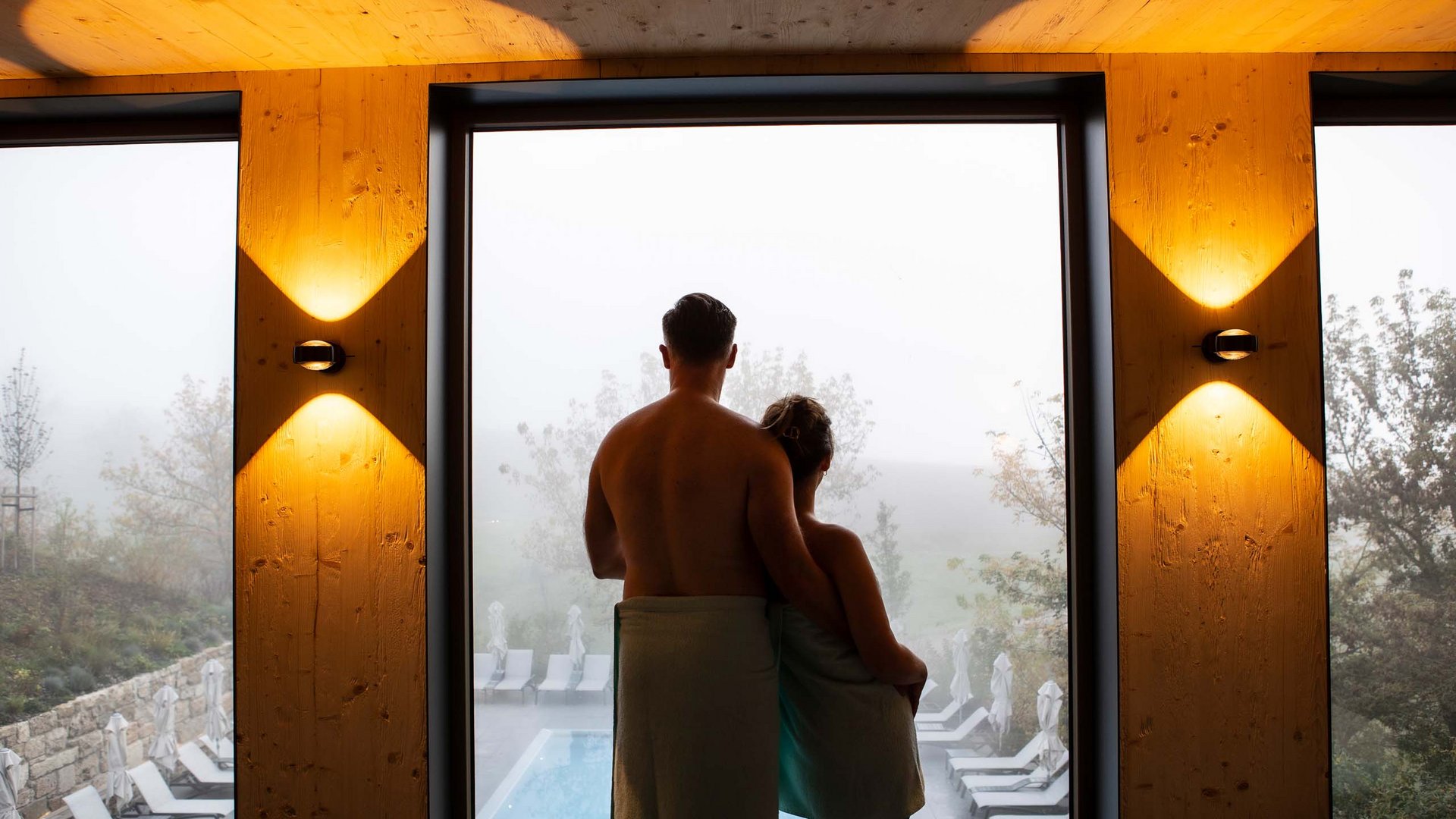 Couple wrapped in towels looking at pool and landscape through large window