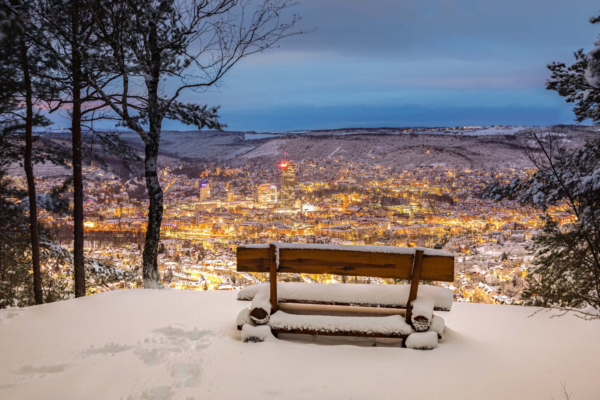 Snow-covered bench overlooking a lit city in a snowy valley at dusk