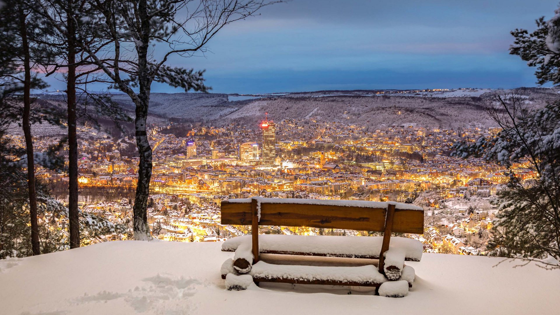 Snow-covered bench overlooking a lit city in a snowy valley at dusk