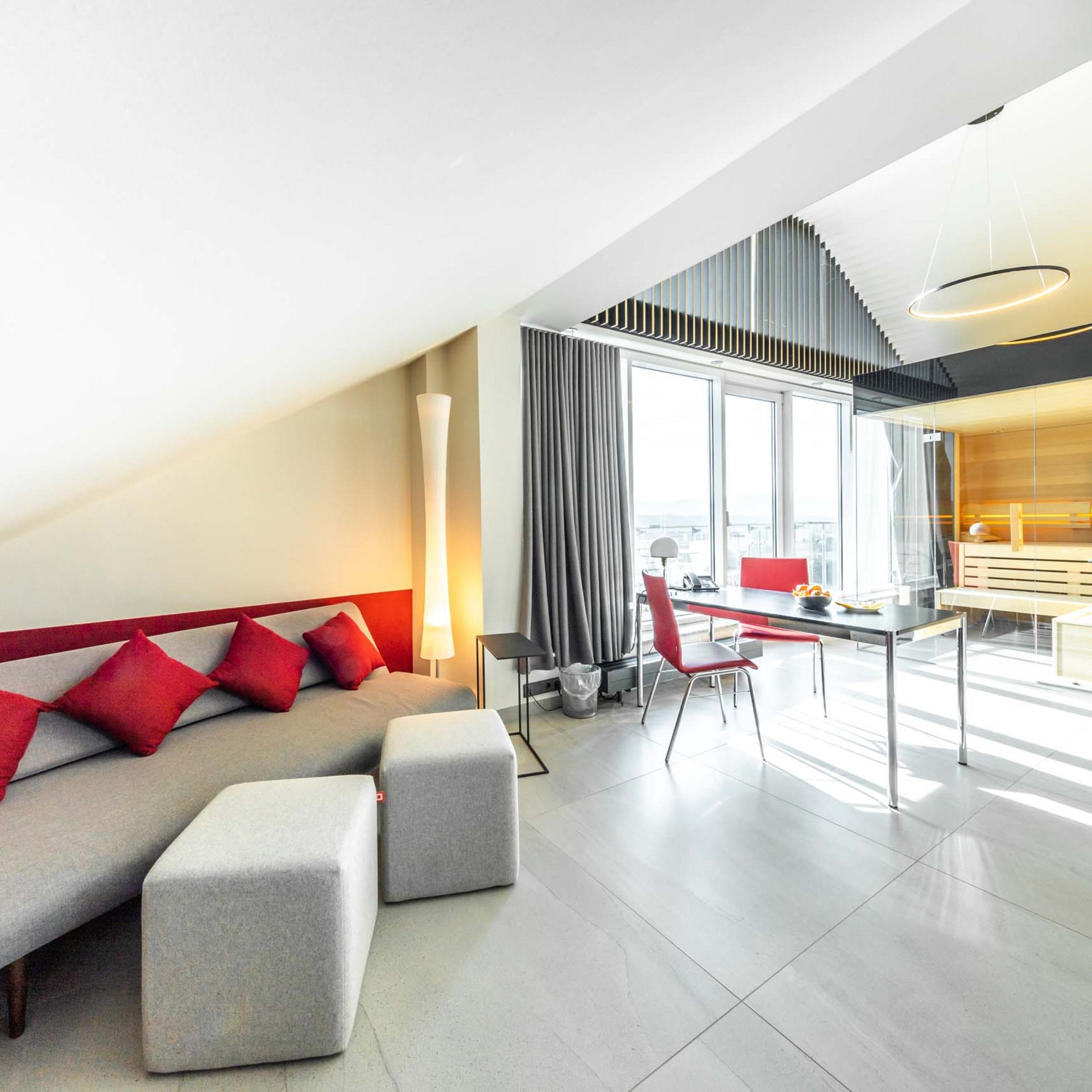 Modernes Wohnzimmer mit grauem Sofa, roten Kissen und Glaswand zur Sauna